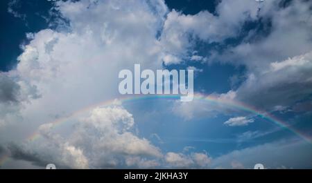 A low angle shot of a long rainbow under a bright blue cloudy sky Stock Photo