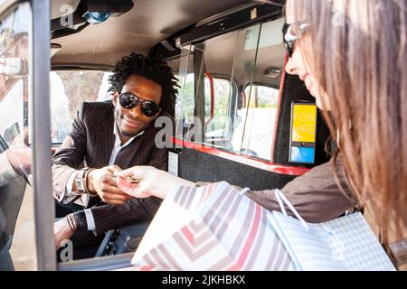 Clients de Londres ; course en taxi. Une jeune femme paie son chauffeur de taxi londonien après une journée de shopping. D'une série d'images avec les mêmes caractères. Banque D'Images