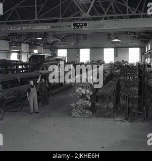 1950s, historique, piles de tiges d'acier dans un grand hangar ou cintre, Port Talbot, pays de Galles, Royaume-Uni. Deux travailleurs de l'acier utilisant des engins de levage aériens fabriqués par Herbert Morris Ltd de Loughborough - des fabricants spécialisés de grues et de blocs de poulie - pour soulever et manœuvrer les tuyaux. Pas de chapeaux de sécurité, juste des casquettes en tissu! Banque D'Images