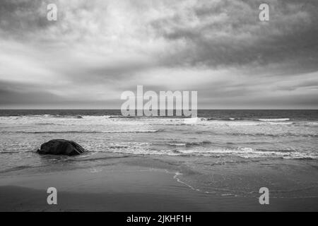 A grayscale shot of a tranquil sandy beach with waves washing the coast Stock Photo