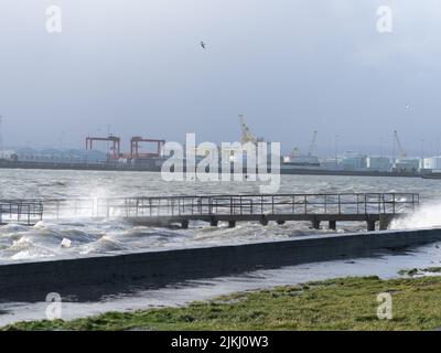 A beautiful shot of pier in the middle of stormy sea with cranes in the port in the background against cloudy sky Stock Photo