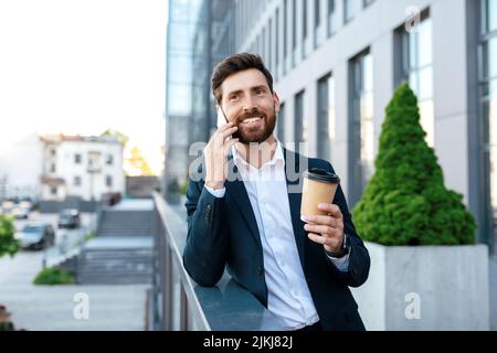 Joyeux homme européen millénaire avec barbe en costume, appel par smartphone, boissons à emporter sur le balcon Banque D'Images