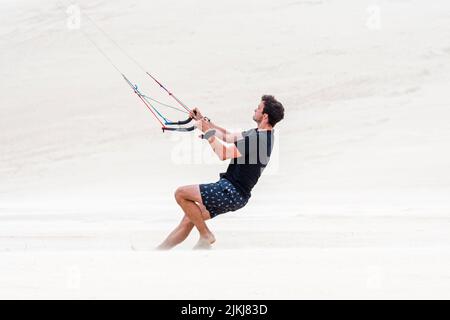 Jeune homme manipulant deux poignées de contrôle d'un entraîneur de quatre lignes de vol parafoil / 4 ligne stunt kite sur la plage de sable dans le vent fort en été Banque D'Images