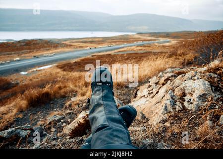 Legs of a man resting in black shoes for mountain tracking against the blurred backdrop of mountains, lake and road during daytime Stock Photo