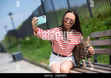 Une jeune femme s'assoit sur un banc avec un chat tabby et prend un selfie sur un smartphone à l'extérieur. Banque D'Images