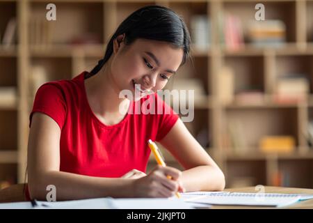 Souriante élégante jeune femme chinoise étudiant en train de faire ses devoirs, se préparant à l'examen seul dans la chambre intérieure, gros plan Banque D'Images