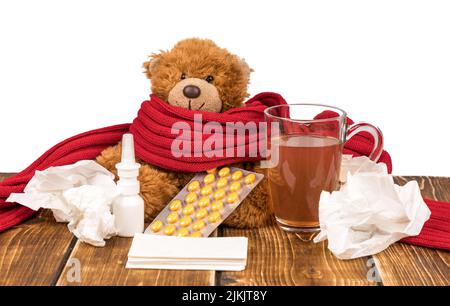 Un ours en peluche avec un spray nasal, des pilules, une écharpe rouge, du thé et des mouchoirs sur une surface en bois - souffrant de maladie Banque D'Images