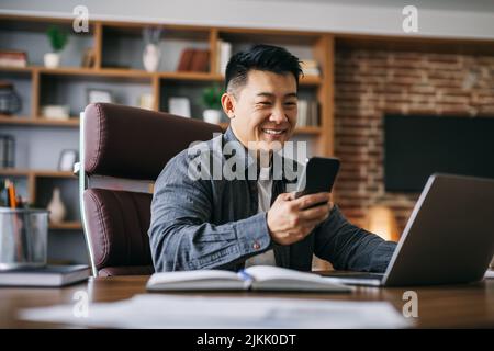 Un homme d'affaires asiatique mûr et heureux qui tape sur son smartphone sur son lieu de travail, à la maison, sur son ordinateur portable Banque D'Images