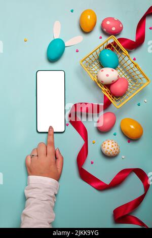 Vue de dessus d'un smartphone adulte avec des œufs de Pâques colorés, un ruban rouge et un petit panier jaune sur fond bleu clair Banque D'Images
