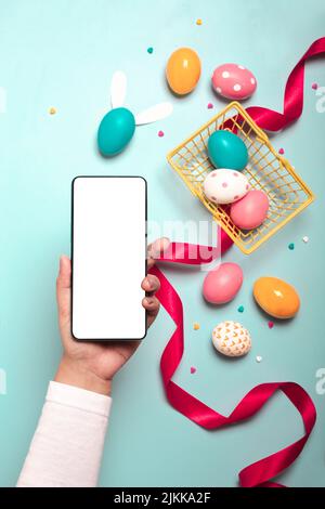 A top view of an adult hand holding smartphone with colorful Easter eggs, red ribbon and small yellow basket on a light blue background Banque D'Images