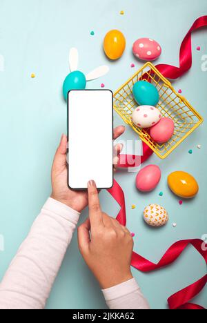 Une vue de dessus des mains adultes tenant un smartphone avec des œufs de Pâques colorés, un ruban rouge et un petit panier jaune sur un fond bleu clair Banque D'Images