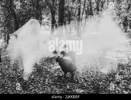 Photo en niveaux de gris d'un homme portant un noir complet assis sur le sol avec un brouillard blanc devant lui au milieu d'une forêt Banque D'Images