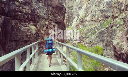 A shot of a woman holding a big backpack and hiking on bridge through the bottom of Grand Canyon, USA Stock Photo