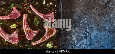 Vue du dessus panorama de côtelettes d'agneau crues placées sur une plaque de cuisson noire en métal avec des verts sur fond sombre dans la cuisine Banque D'Images