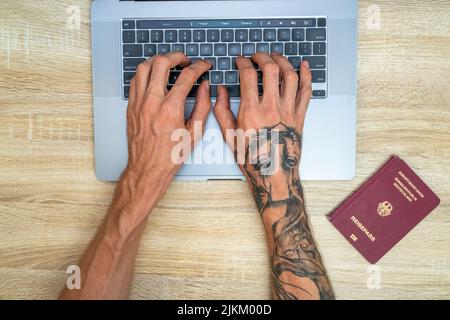 Vue de dessus des mains tatouées tapant sur un ordinateur portable à la recherche d'un vol, avec un passeport allemand sur le côté Banque D'Images