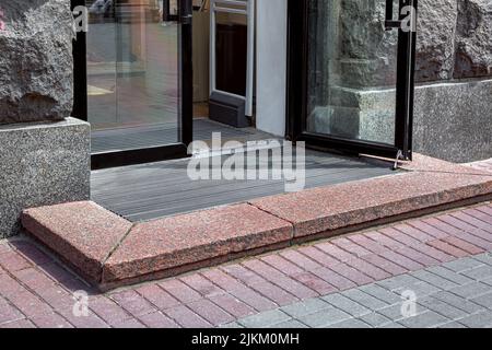 entrée avec seuil de granit pour stocker depuis le trottoir piétonnier de la plieuse sur le tapis de pied à travers la porte en verre avec cadre noir, façade en pierre rugueuse d'ex Banque D'Images