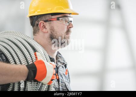 Caucasien électricien Entrepreneur dans ses années 30 avec mur métal aluminium conduit flexible sur son épaule.Installation de tuyaux électriques métalliques.Construction Banque D'Images