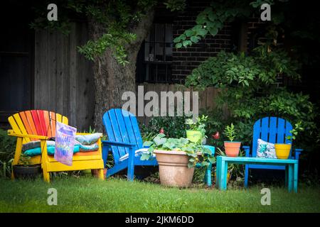 Salon extérieur coloré - chaises Adirondack peintes avec pots de plantes sous l'arbre dans un endroit ombragé et drapeau de la maison Sweet Home Banque D'Images