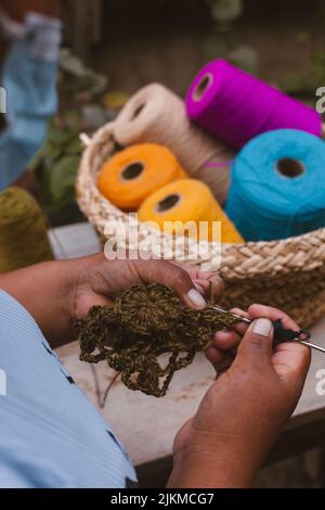 A vertical shot of the hands knitting a decoration and colorful threads in a basket in the background Stock Photo