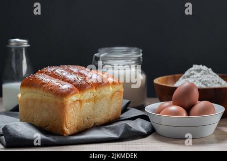 A closeup shot of a freshly baked milk bread accompanied with flour, eggs, sugar, and milk Stock Photo