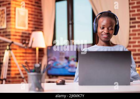Jeune femme afro-américaine dans un casque regardant des vidéos sur un ordinateur portable à la maison. Femme souriante étudiant à l'université de cours à distance, studyin avec ordinateur dans la salle moderne Banque D'Images