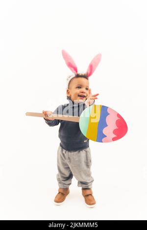 Tout-petit afro-américain très mignon et plein de plaisir avec des oreilles de lapin de pâques tenant un gros œuf de pâques sur le bâton - isolé. Photo de haute qualité Banque D'Images