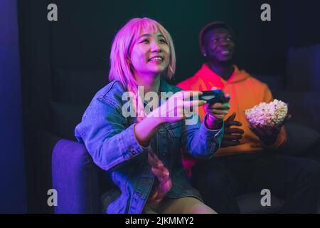 Fille asiatique avec des cheveux roses souriant jouant jeu de console à côté de son ami mangeant pop-corn assis sur le canapé ensemble. Éclairage bleu. Photo de haute qualité Banque D'Images