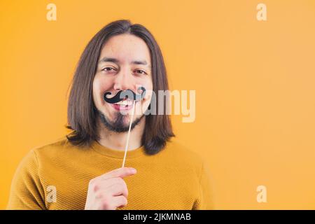 Jeune latin à poils longs barbu dans sa fête de 30s et portant une moustache en papier noir dans une pièce. Photo de haute qualité Banque D'Images