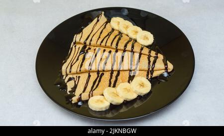 Un gros plan de crêpes au sirop de chocolat et aux bananes sur une assiette noire Banque D'Images