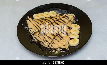 Un gros plan de crêpes au sirop de chocolat et aux bananes sur une assiette noire Banque D'Images