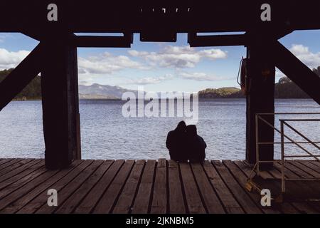 Un couple silhoueté de dos assis sur des planches de bois plancher sous un pont regardant la mer d'eau Banque D'Images