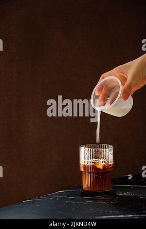 A closeup shot of a man pouring milk into a glass cup of coffee on blurred background Banque D'Images