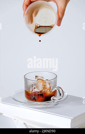 Un cliché vertical d'une main ajoutant progressivement du café à une tasse avec des glaçons sur fond blanc Banque D'Images