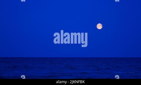 The rising luminous full moon over the dark blue Baltic Sea. The luminous moon forms a strong contrast to the blue sky. Stock Photo