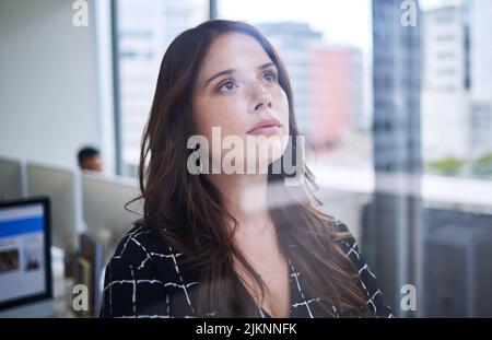 Ayez le courage de poursuivre vos rêves. Une jeune femme d'affaires regardant réfléchie tout en se tenant debout à une fenêtre dans un bureau. Banque D'Images