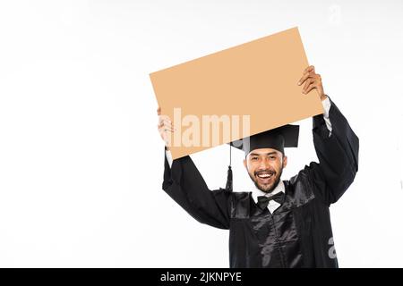 Riant étudiant diplômé de sexe masculin portant tosa tenant un espace en carton vierge Banque D'Images