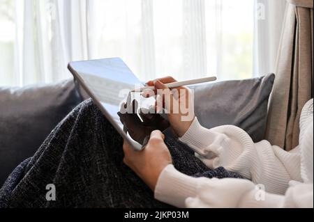 Une jolie femme se détend dans le salon, se repose sur un canapé confortable et utilise un pavé tactile numérique pour tablette. Banque D'Images