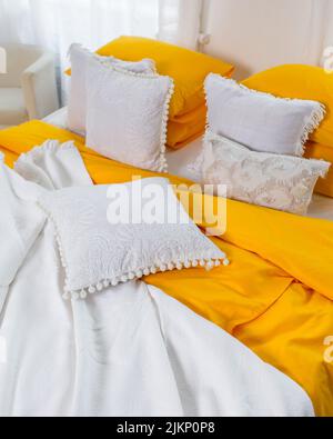 A bright vertical shot of a bed with white and yellow pillows, bedsheets, and covers in a white room Stock Photo