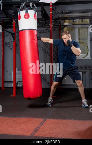 Jeune homme barbu, combattant et entraîneur d'arts martiaux mixtes, pratique des coups de poing avec sa main droite sur le sac de poinçonnage rouge dans la salle de sport. Banque D'Images