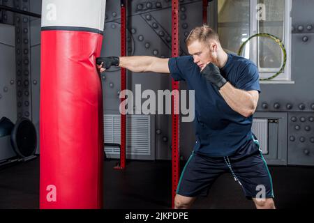 Jeune homme barbu, combattant et entraîneur d'arts martiaux mixtes, pratique des coups de poing avec sa main droite sur le sac de poinçonnage rouge dans la salle de sport. Banque D'Images