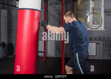 Jeune homme barbu fort, combattant et entraîneur d'arts martiaux mixtes, s'entraîne dans la salle de gym. Frappe avec sa main gauche sur un sac de poinçonnage rouge. Banque D'Images