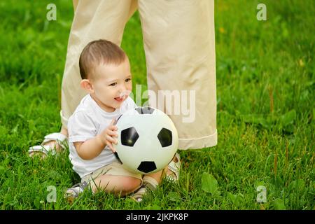 Bébé garçon et mère heureux avec un ballon de football sur l'herbe verte. Un enfant souriant en vêtements blancs est assis avec une balle dans ses mains, un an Banque D'Images