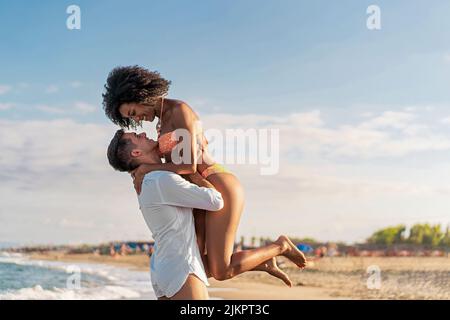 Couple multiethnique de jeunes amoureux sur la plage - le gars ramasse sa petite amie afro-américaine sur fond de ciel bleu - peop Banque D'Images