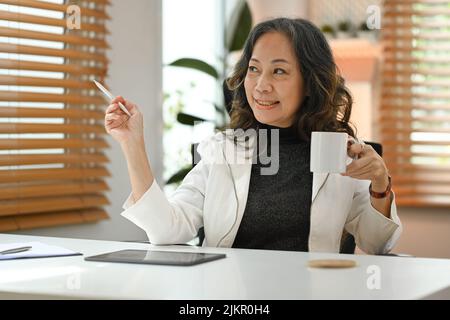 Femme d'affaires satisfaite et mûre tenant une tasse de café et regardant par la fenêtre, profitez d'une ambiance paisible sans stress Banque D'Images