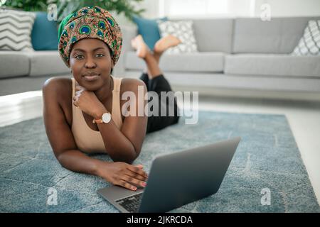 Une jeune femme africaine heureuse assise avec ses jambes croisées sur son tapis à la maison à l'aide d'un ordinateur portable et regardant dans l'appareil photo Banque D'Images