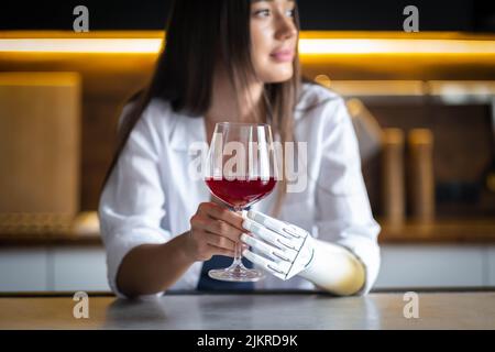 Une jeune fille calme et réfléchie avec un bras prothétique tient un verre de vin à la maison, une femme handicapée mène à la vie normale un membre prothétique artificiel, les gens avec Banque D'Images