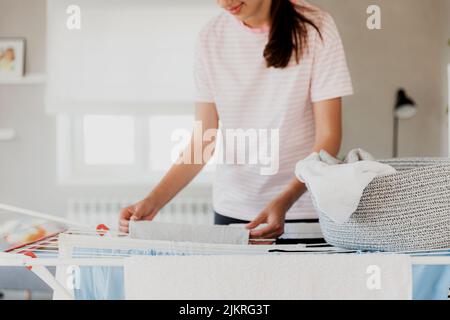 Bonne jeune brune femme caucasienne souriant, pendant des vêtements propres et humides sur le sèche-linge à la maison. Femme au foyer les mains de gros plan en étalant le linge du panier. Une fille met des serviettes après les avoir séchées Banque D'Images