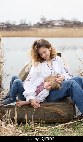 triste petit garçon et jeune belle femme dans les vêtements nationaux brodés se reposent sur le rivage du lac. Famille, réfugiés, unité, soutien, soins. Ukraine Banque D'Images