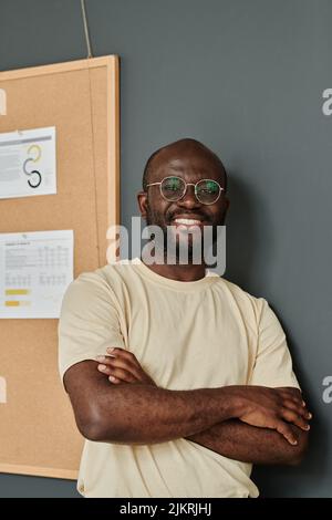 Portrait d'un homme d'affaires africain réussi en lunettes debout avec ses bras croisés au bureau et souriant Banque D'Images