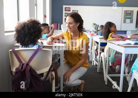 Une jeune enseignante caucasienne souriante parle à un garçon afro-américain assis à son bureau en classe Banque D'Images